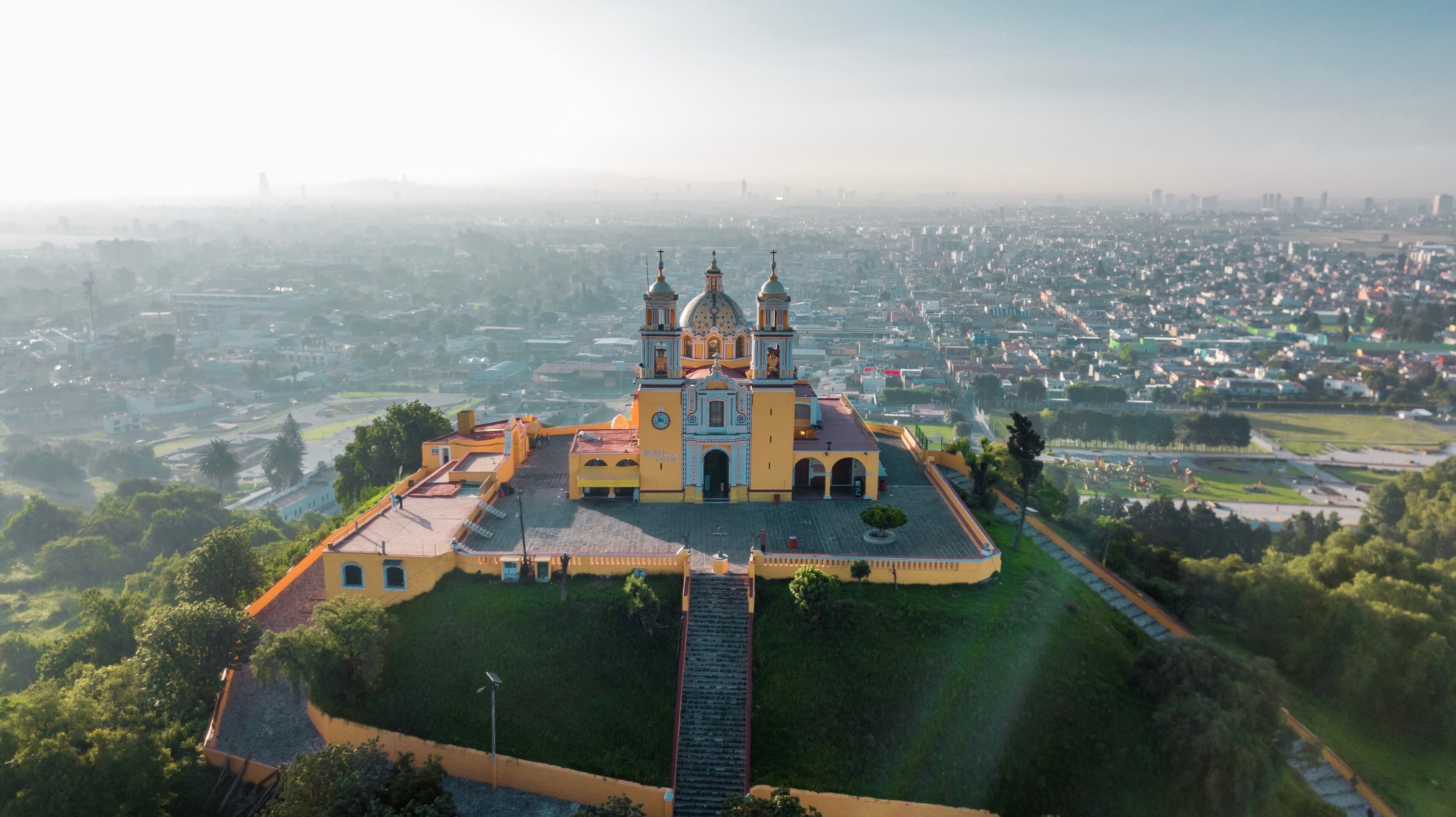 An aerial view of a large, ornate church with two towers, surrounded by a well-maintained park and a cityscape in the background.