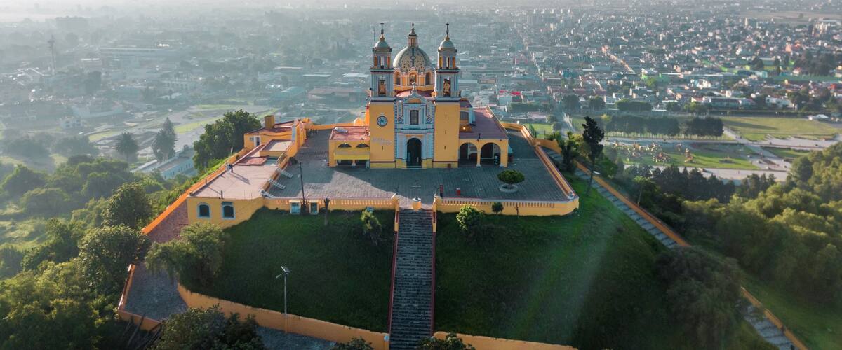 An aerial view of a large, ornate church with two towers, surrounded by a well-maintained park and a cityscape in the background.