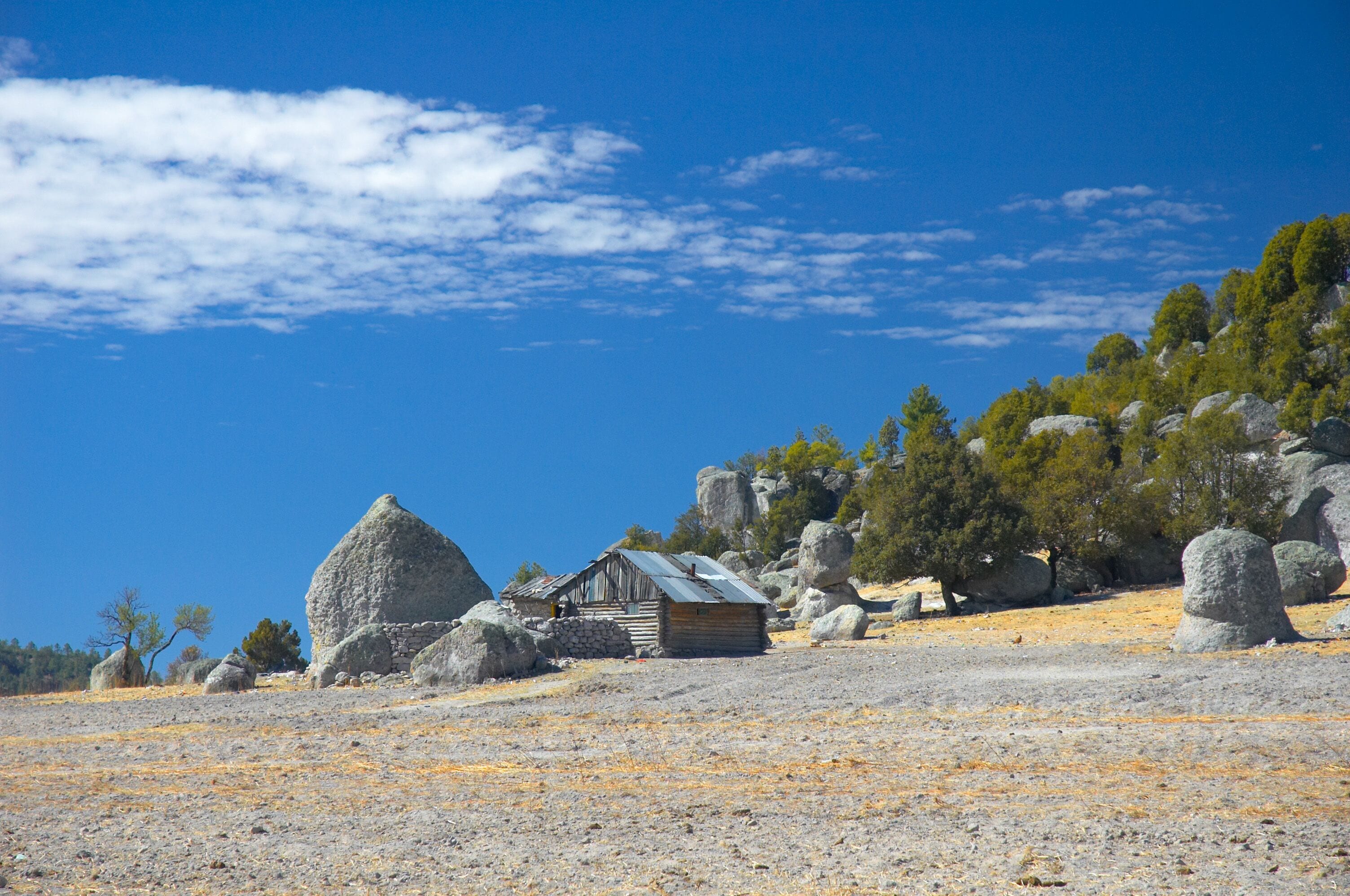 Un paisaje rocoso con una pequeña cabaña de madera, grandes rocas y vegetación escasa bajo un cielo azul despejado.