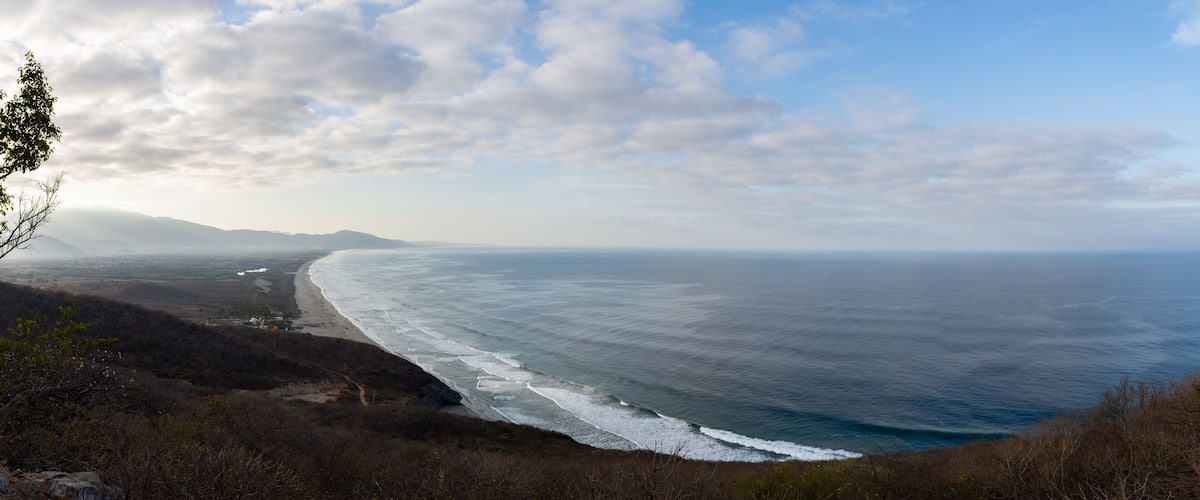 A coastal landscape with a beach, ocean waves, and distant mountains under a partly cloudy sky.