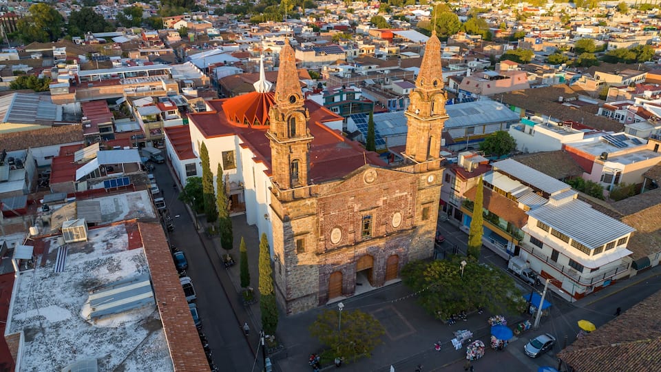 A historic church with two towers, surrounded by a densely packed urban area with various buildings and a mountainous backdrop.