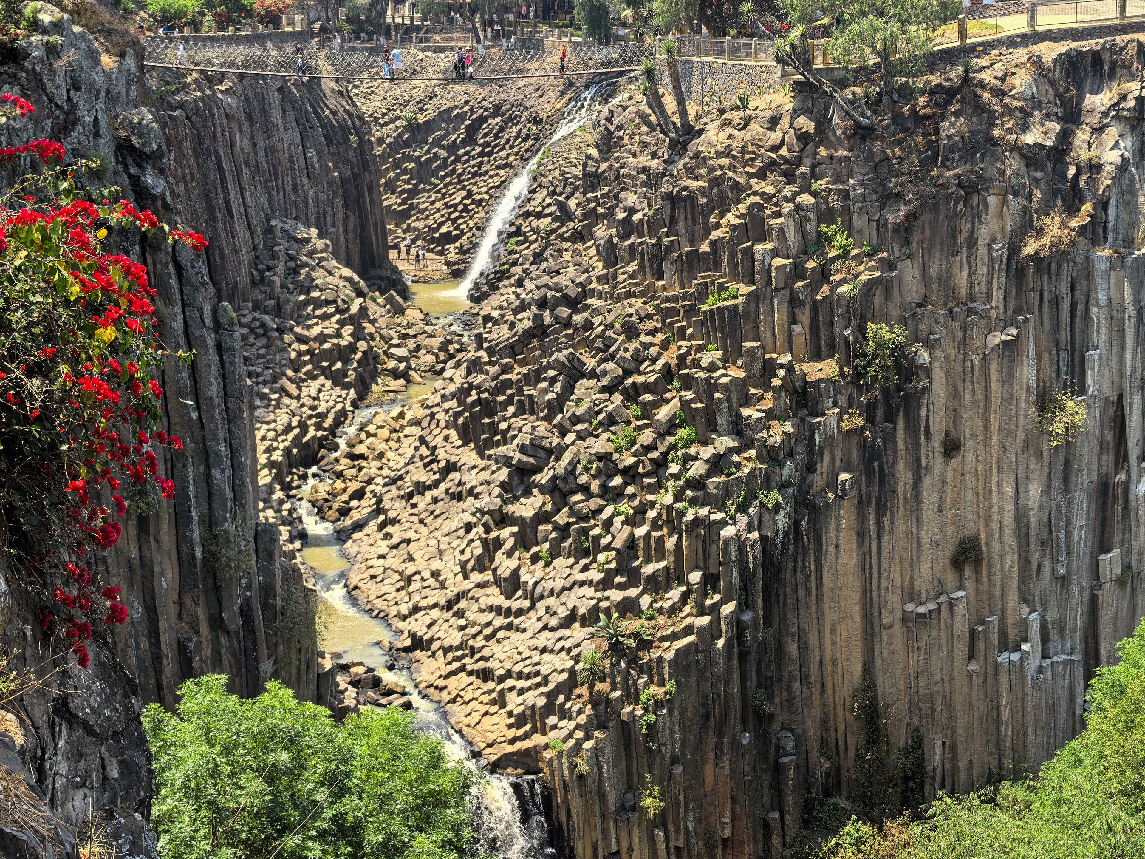 A waterfall cascading down a rocky gorge with a wooden bridge overhead.