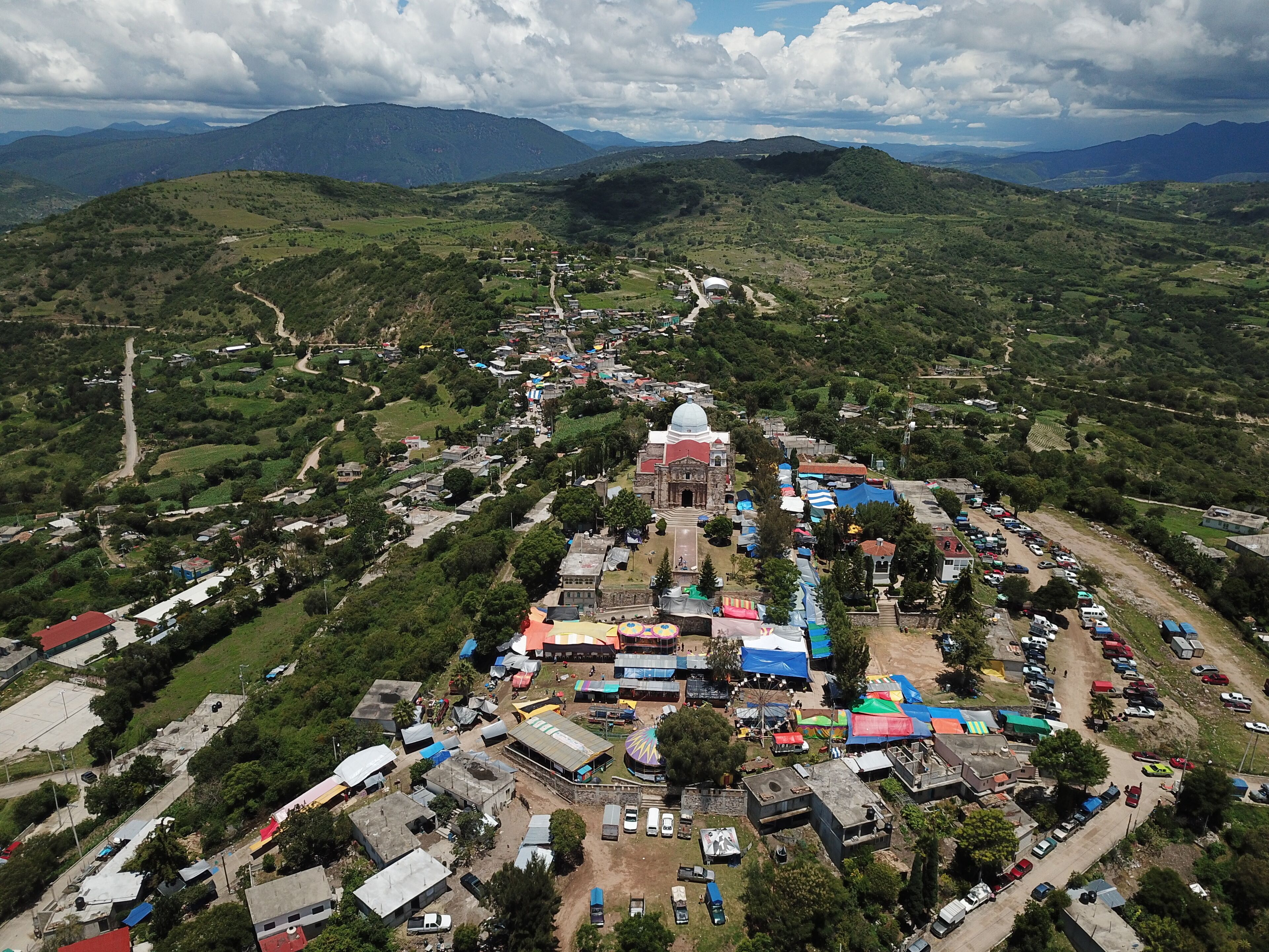 An aerial view of a town with a prominent church, surrounded by lush green hills and mountains.