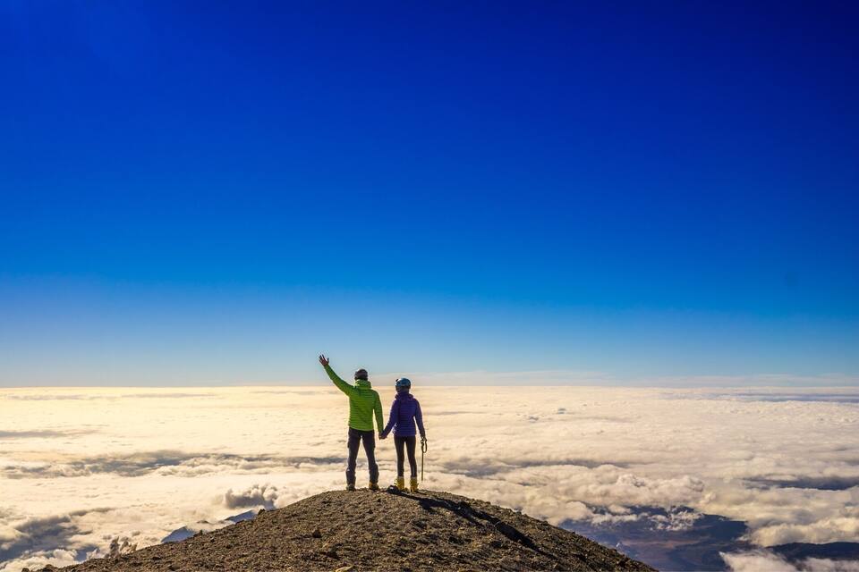Two people standing on a mountain peak with a clear blue sky and clouds below.