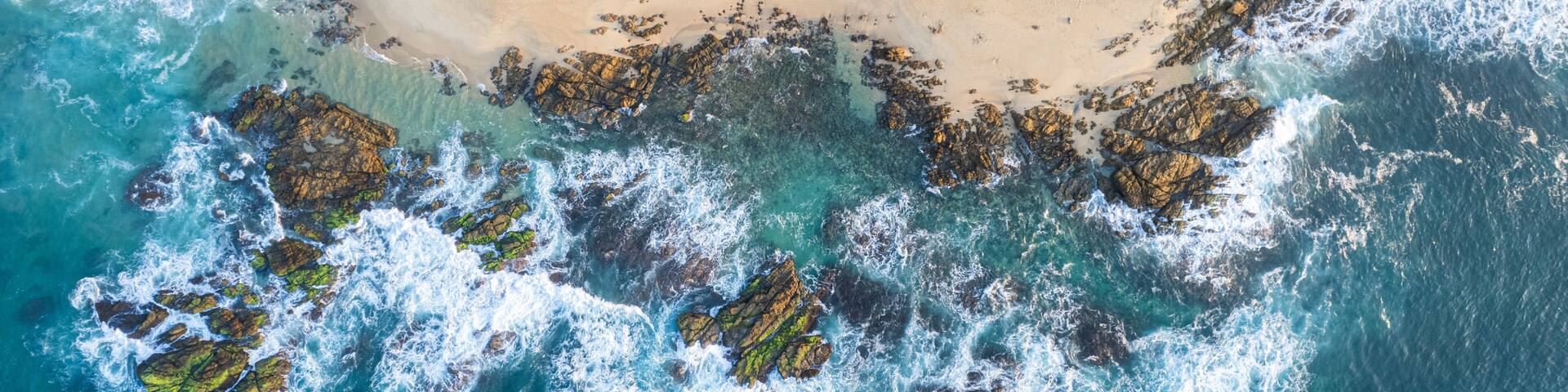 An aerial view of a beach with rocky outcrops and a small settlement with palm trees.