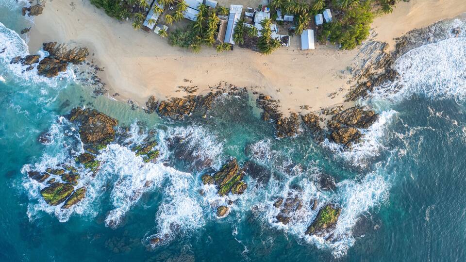An aerial view of a beach with rocky outcrops and a small settlement with palm trees.