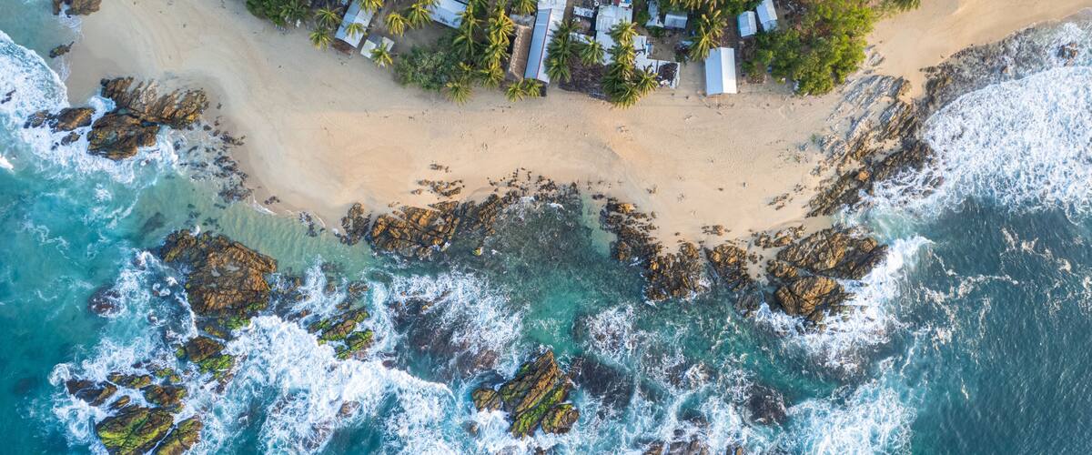 An aerial view of a beach with rocky outcrops and a small settlement with palm trees.