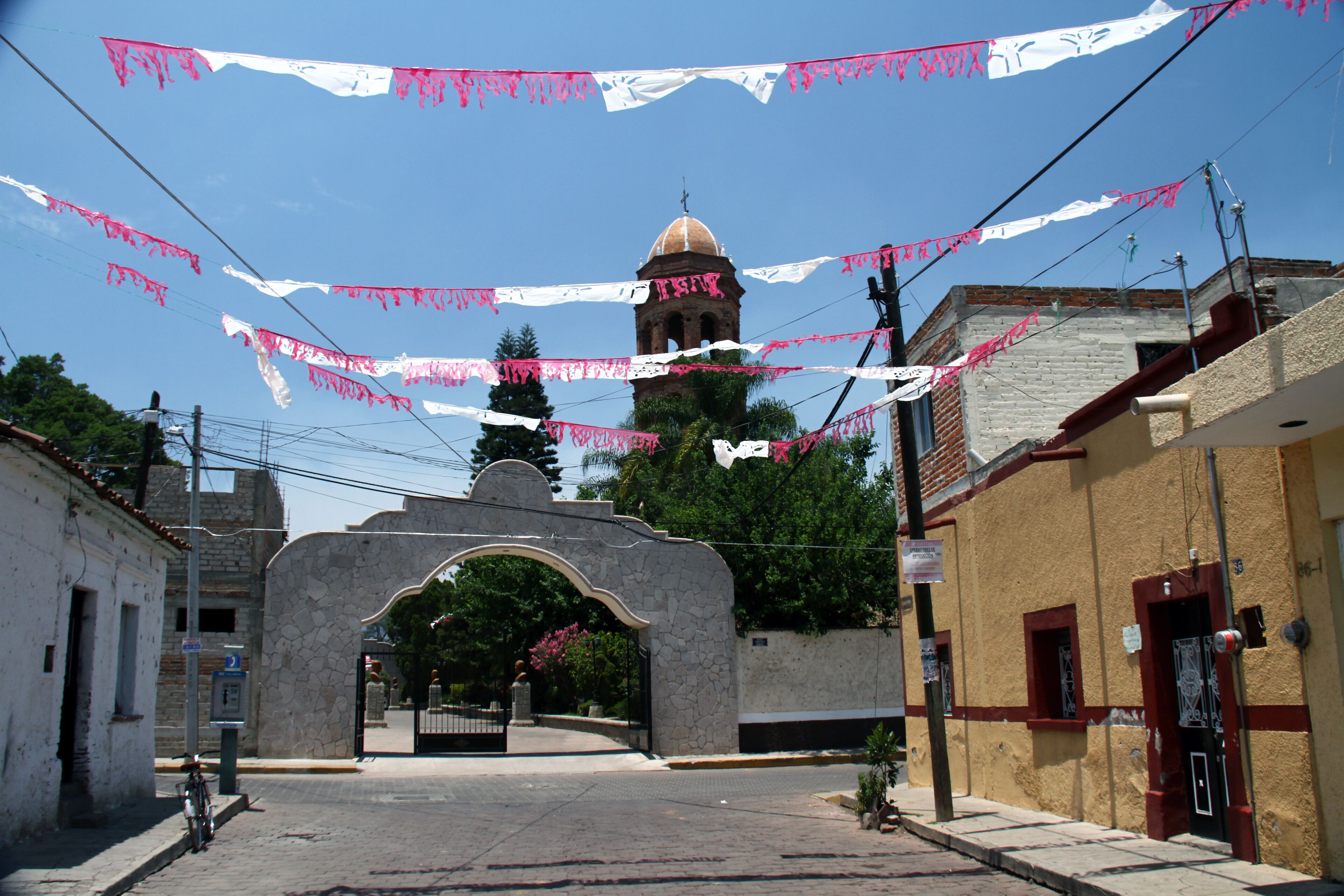 A street with a stone archway, a bell tower, and pink and white banners.