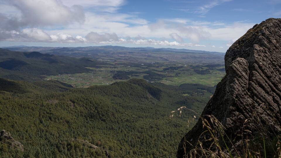 A mountainous landscape with dense forests, a prominent rock formation, and a distant village.