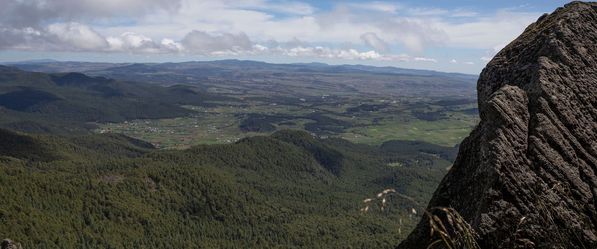 A mountainous landscape with dense forests, a prominent rock formation, and a distant village.