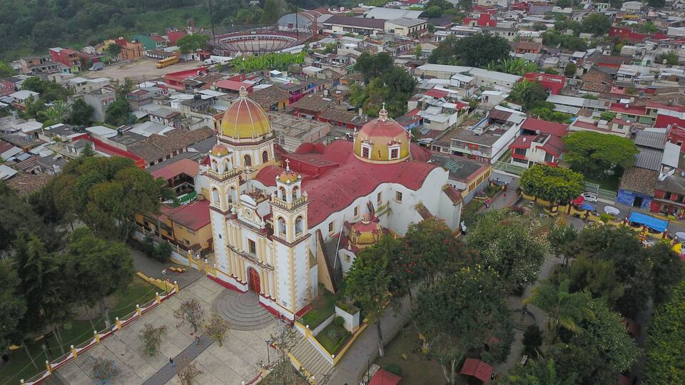 An aerial view of a town with a prominent church featuring a large dome and two smaller domes, surrounded by residential buildings and greenery.