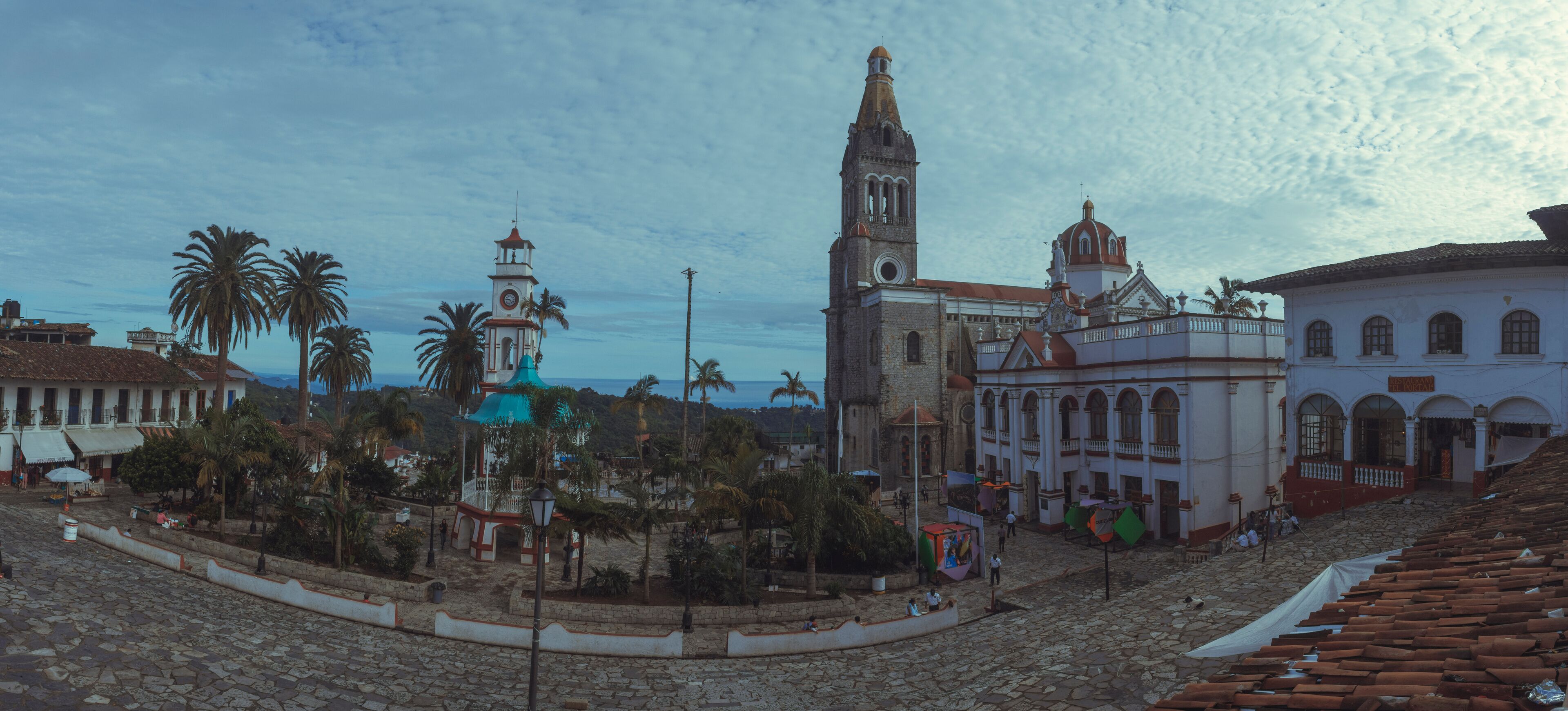 A historic plaza with cobblestone streets, palm trees, and colonial architecture including a church with a bell tower.