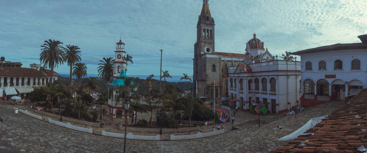 A historic plaza with cobblestone streets, palm trees, and colonial architecture including a church with a bell tower.