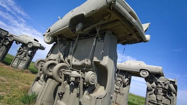 Um veículo militar grande e enferrujado, com a frente destruída, cercado por um campo gramado sob um céu claro.