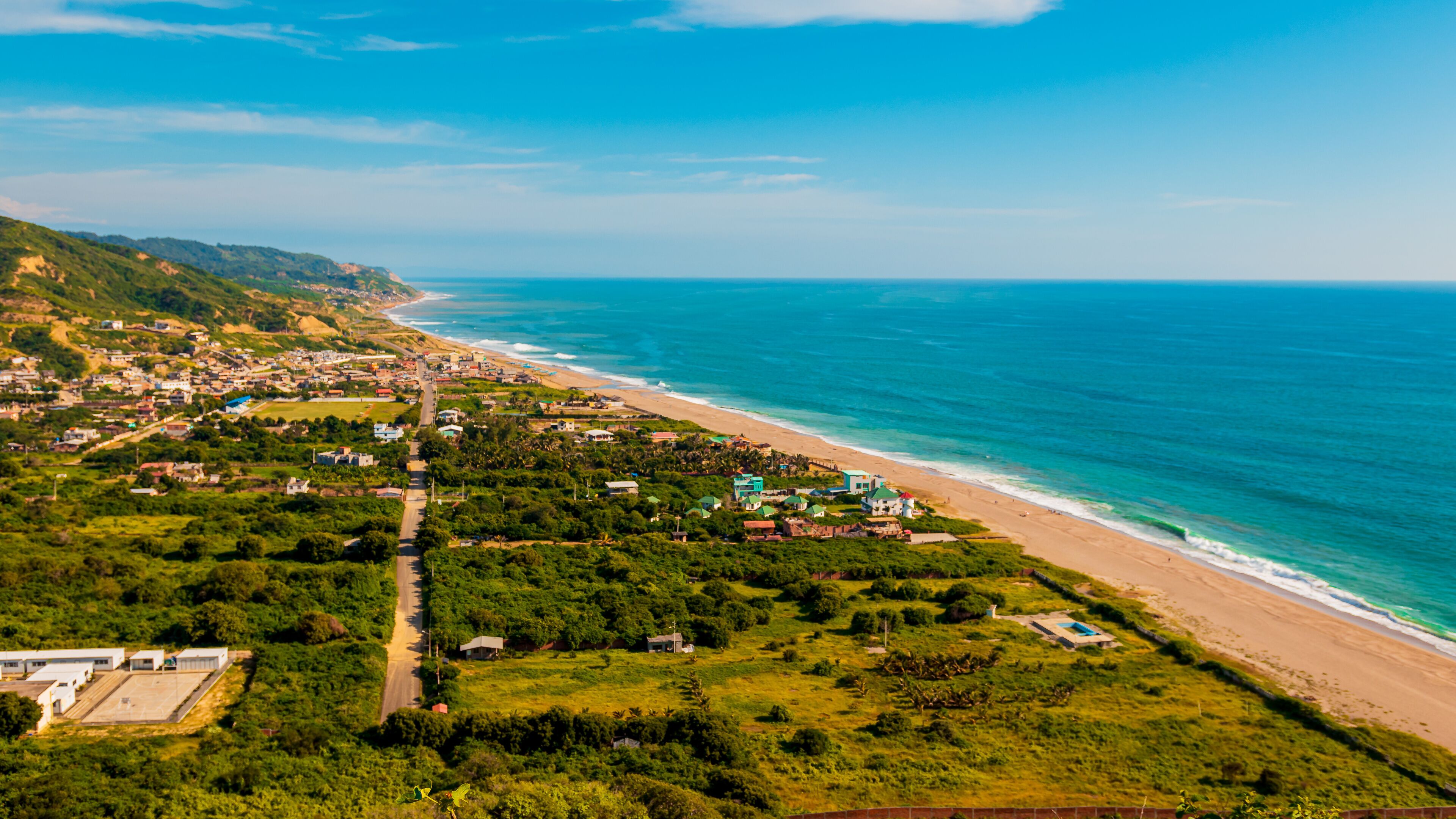 A coastal town with a beach, green hills, and a clear blue sky.