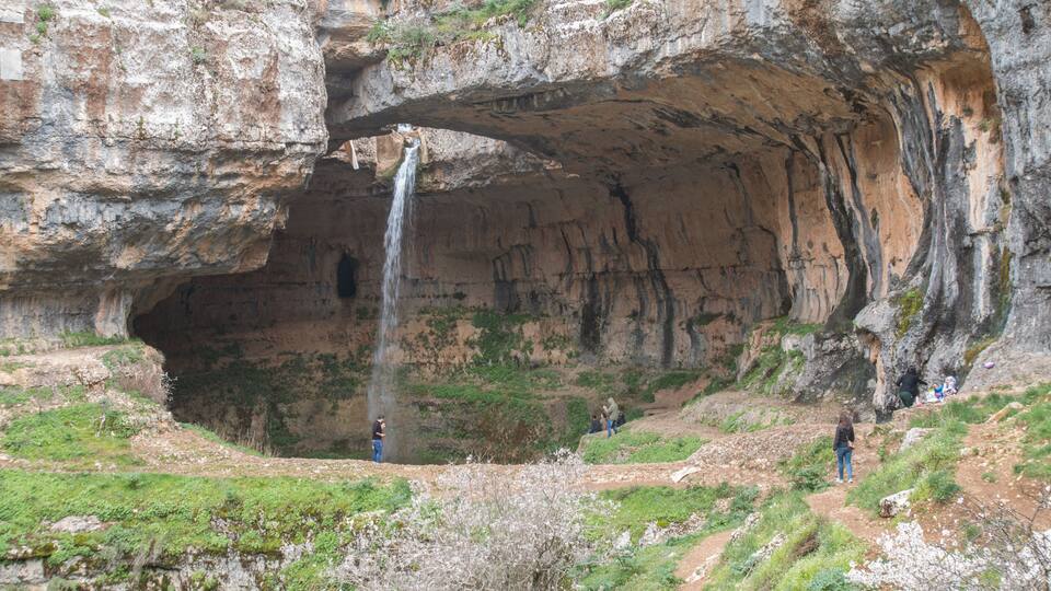 A waterfall cascading through a natural rock arch formation with people observing the scene.
