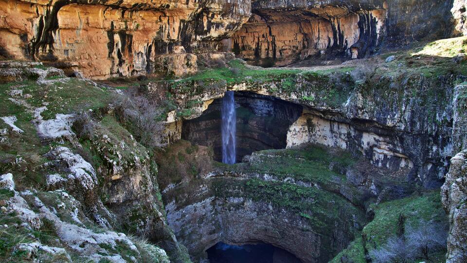 A cave entrance with a waterfall, surrounded by rocky cliffs and greenery.