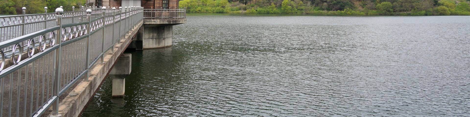 A wooden structure on a bridge over a calm lake, surrounded by lush green hills and mountains.