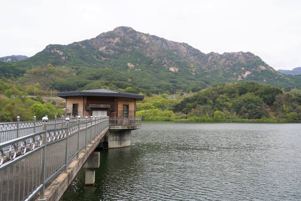 A wooden structure on a bridge over a calm lake, surrounded by lush green hills and mountains.