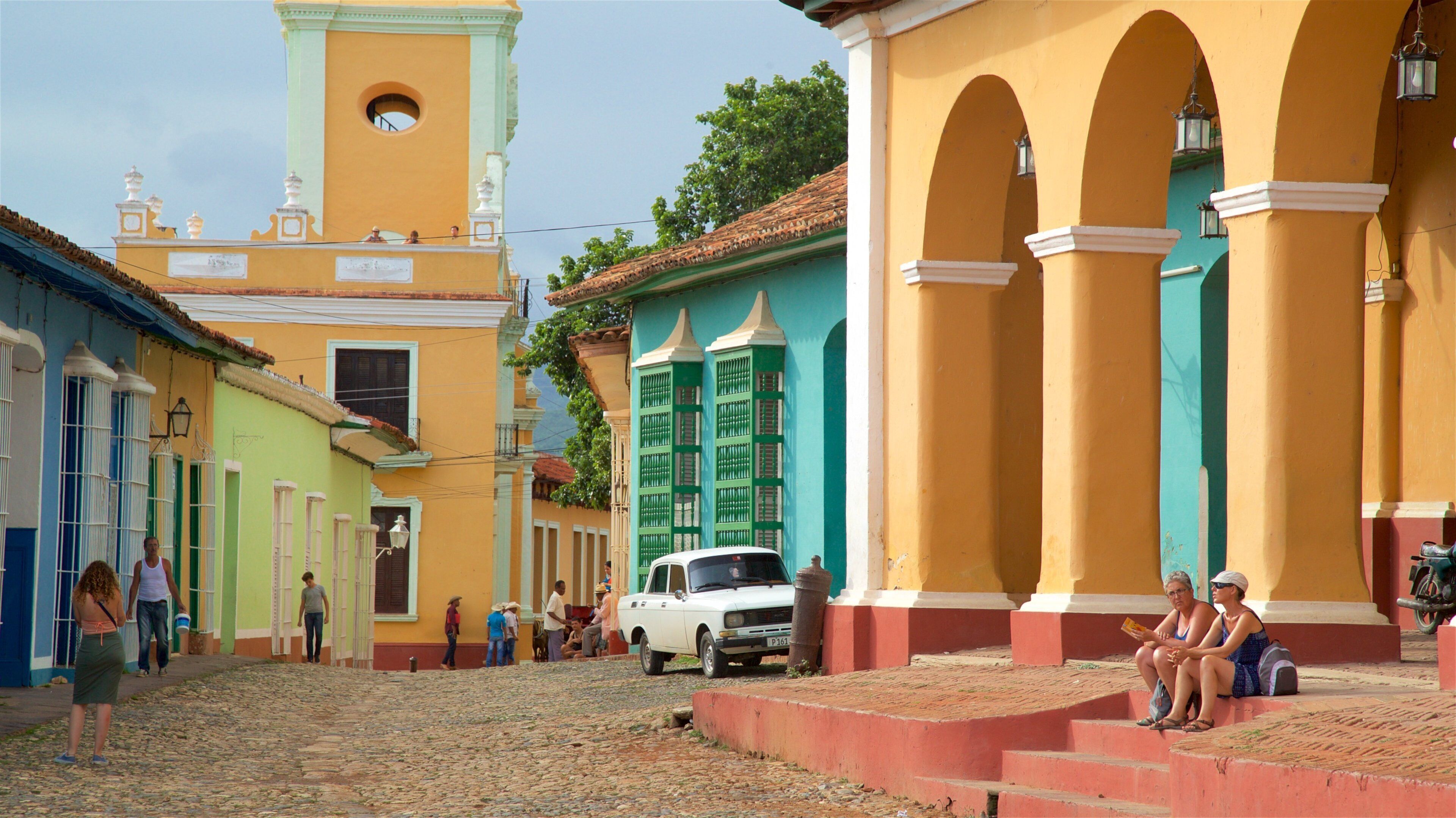 A cobblestone street lined with colorful buildings, a white car parked on the side, and people walking and sitting on the steps.