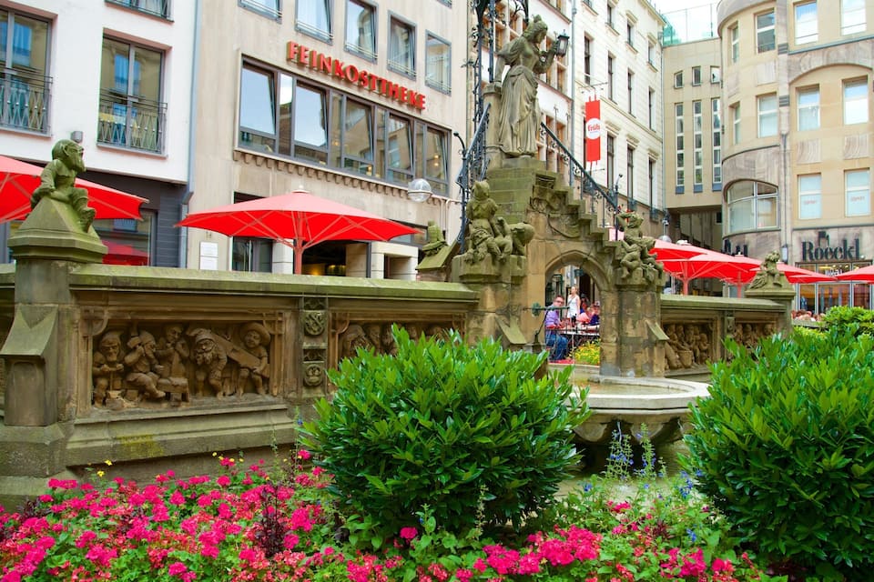 A fountain with sculptures, surrounded by red umbrellas and pink flowers.