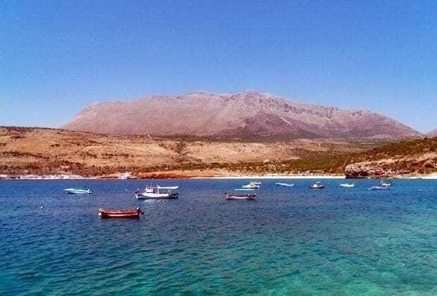 A coastal landscape with boats on clear blue water and a mountain in the background.