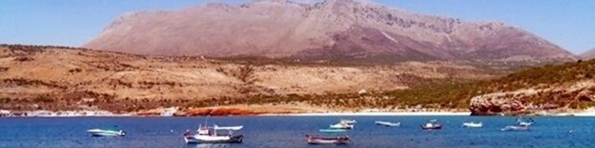 A coastal landscape with boats on clear blue water and a mountain in the background.