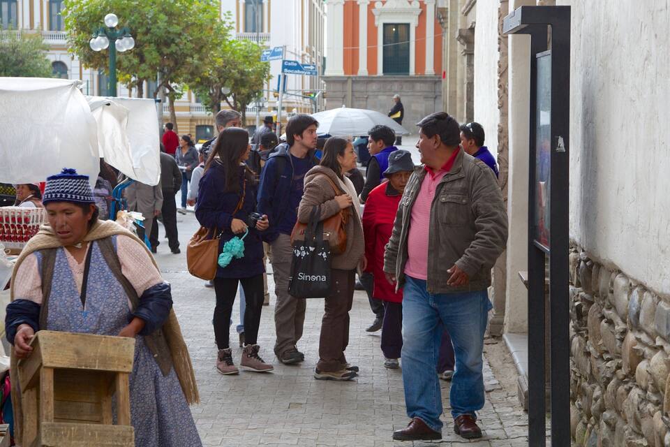Rua movimentada com pessoas caminhando e carregando sacolas. Prédios com colunas e uma placa com a inscrição 'Calle Socabaya'.
