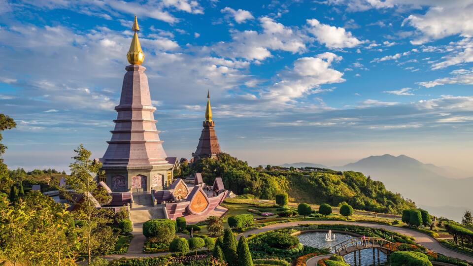 A temple complex with two tall spires, surrounded by lush gardens and a small bridge over a pond.