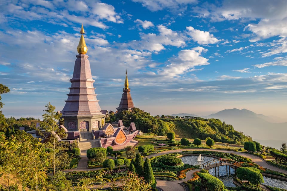 A temple complex with two tall spires, surrounded by lush gardens and a small bridge over a pond.