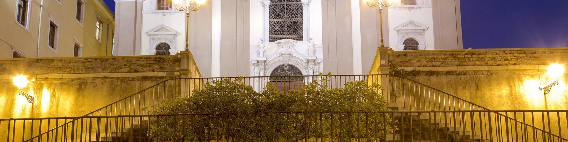 A grand white church with ornate columns and a large arched window, surrounded by a well-lit staircase and a fence.