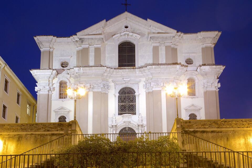 A grand white church with ornate columns and a large arched window, surrounded by a well-lit staircase and a fence.