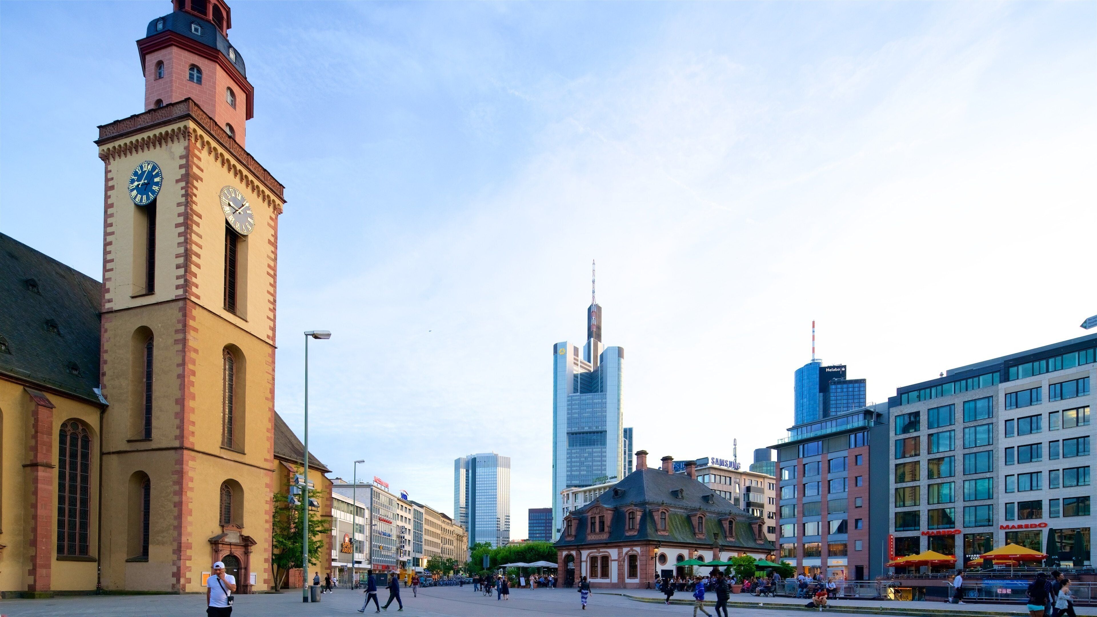 A historic clock tower, modern skyscrapers, and a plaza with people walking.