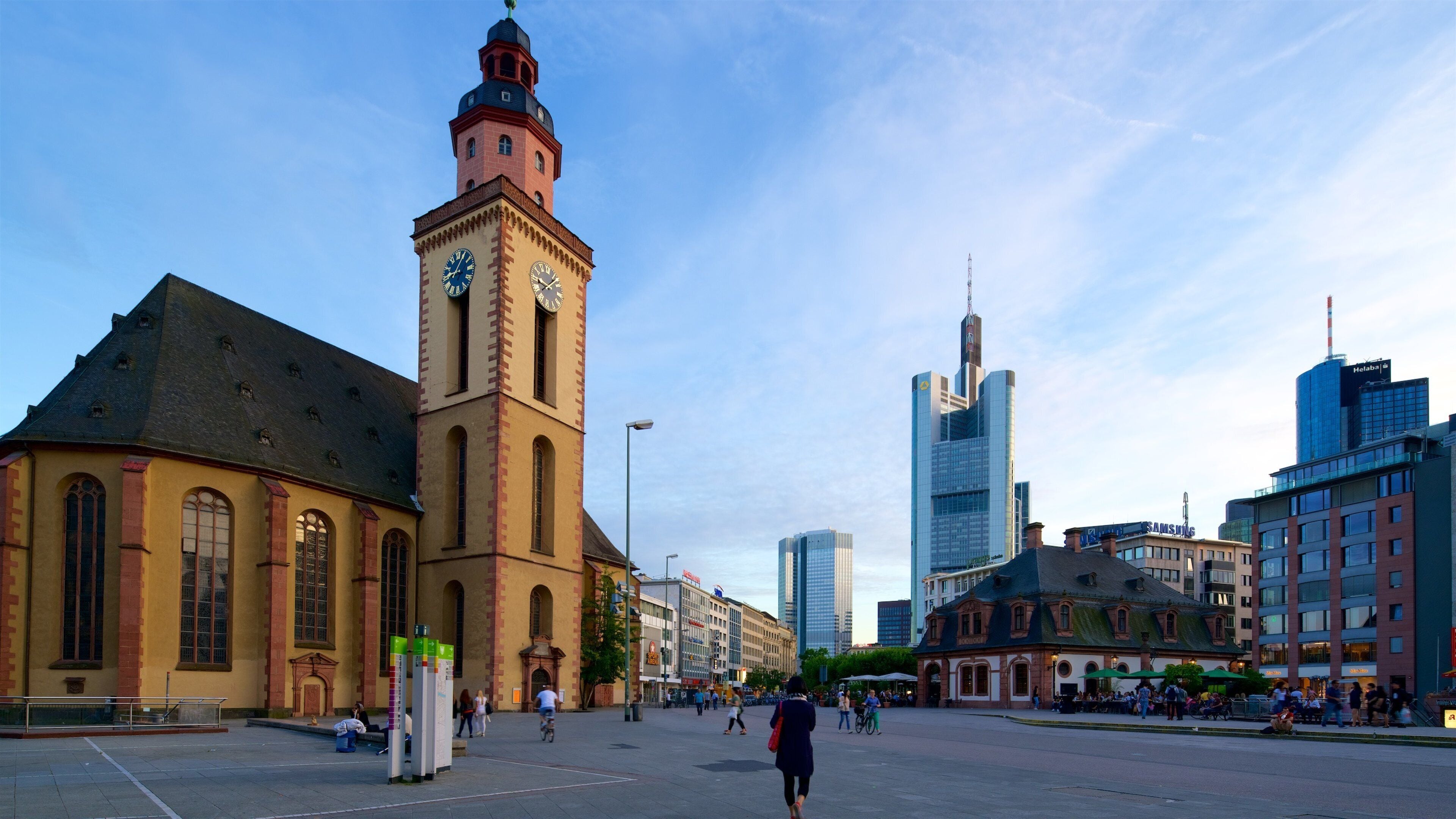 A historic church with a clock tower, surrounded by modern skyscrapers and a bustling square.