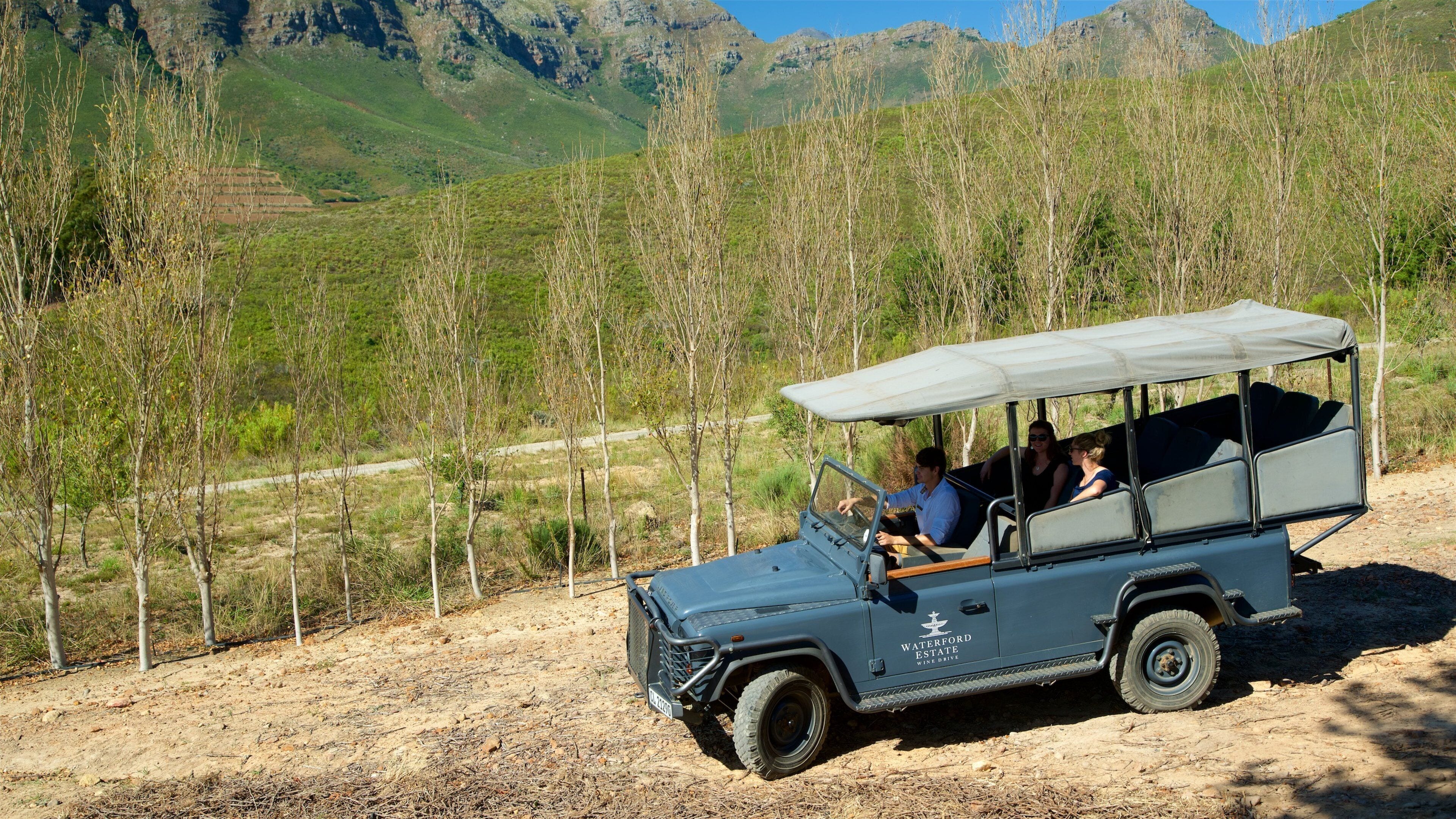 A blue off-road vehicle with a canopy, labeled "Waterford Estate Wine Drive," parked on a dirt road with a mountainous backdrop.