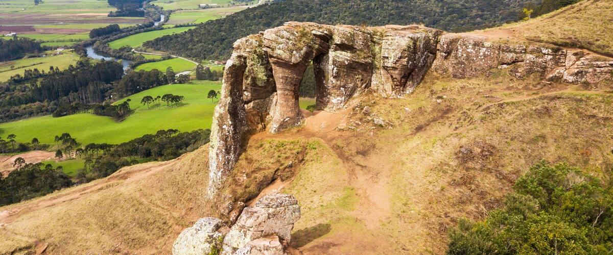 A large rock formation with a natural arch, surrounded by rolling hills and a river in the valley below.
