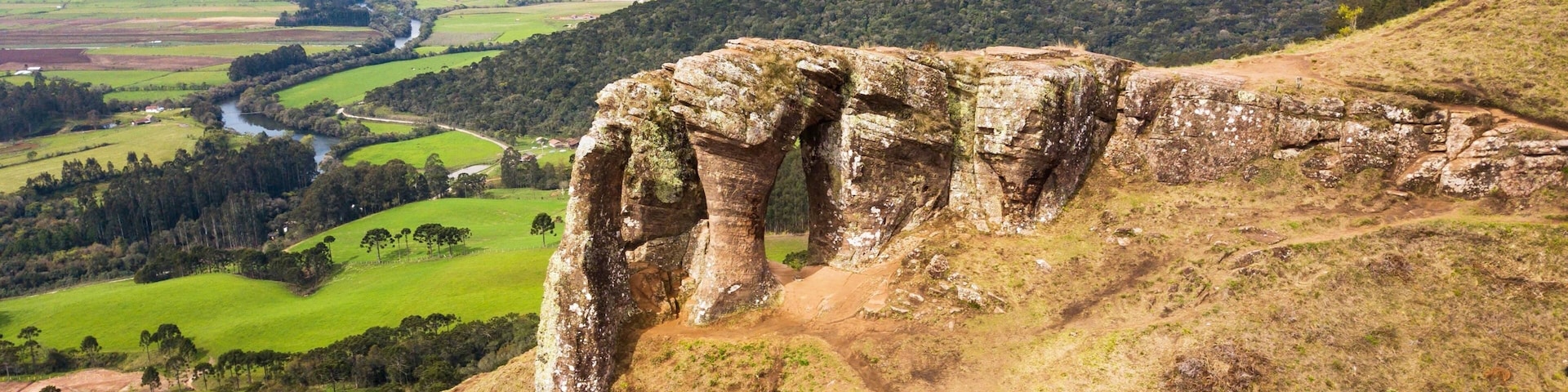 A large rock formation with a natural arch, surrounded by rolling hills and a river in the valley below.