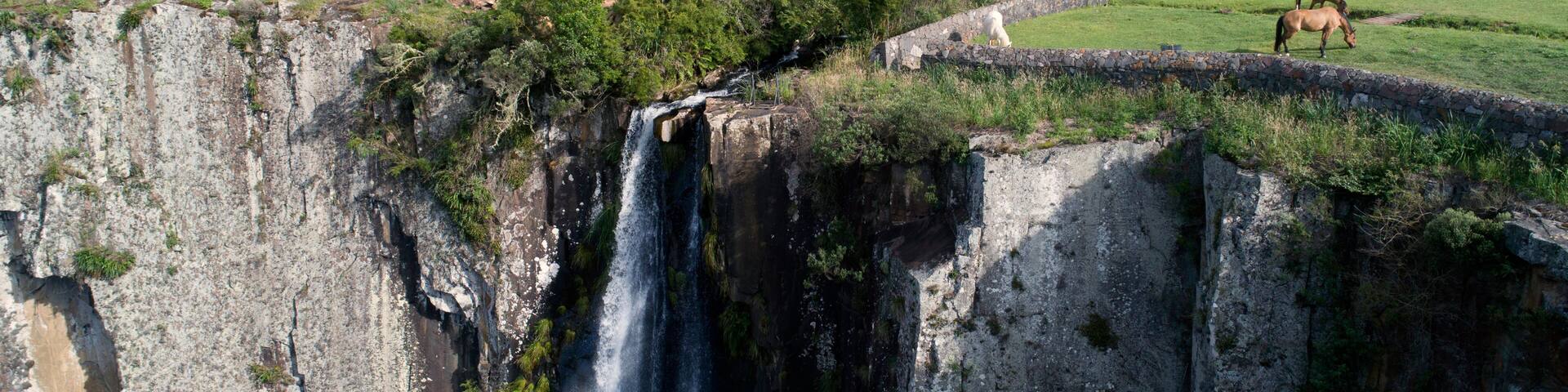 Une cascade, une maison en bois et un cheval qui broute dans un paysage rural.