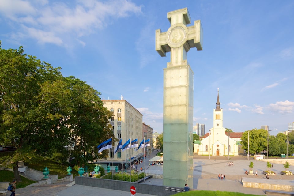 A large cross monument in a city square with surrounding buildings and trees.