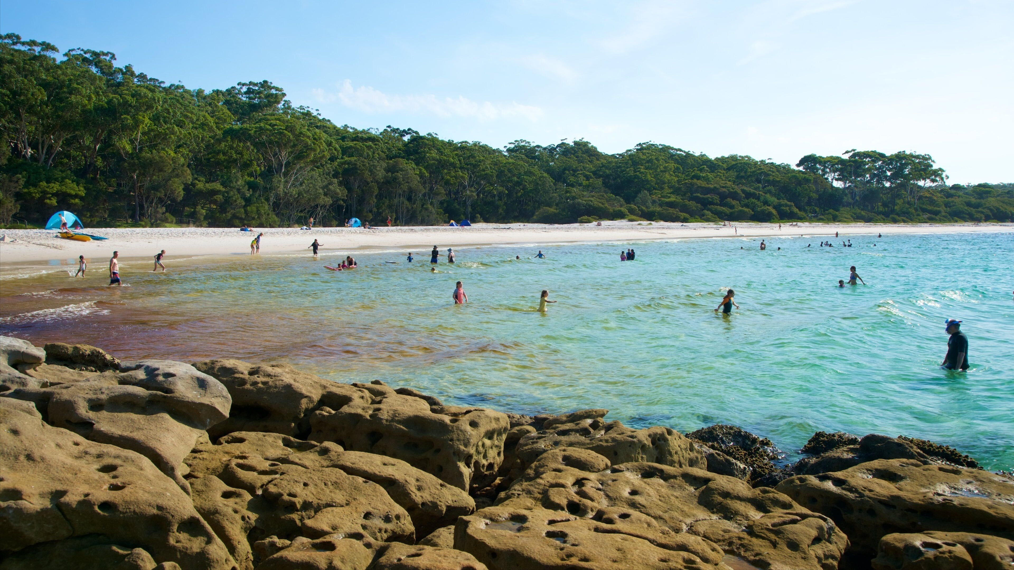 Una playa con agua turquesa cristalina, costa rocosa y gente nadando y tomando el sol.