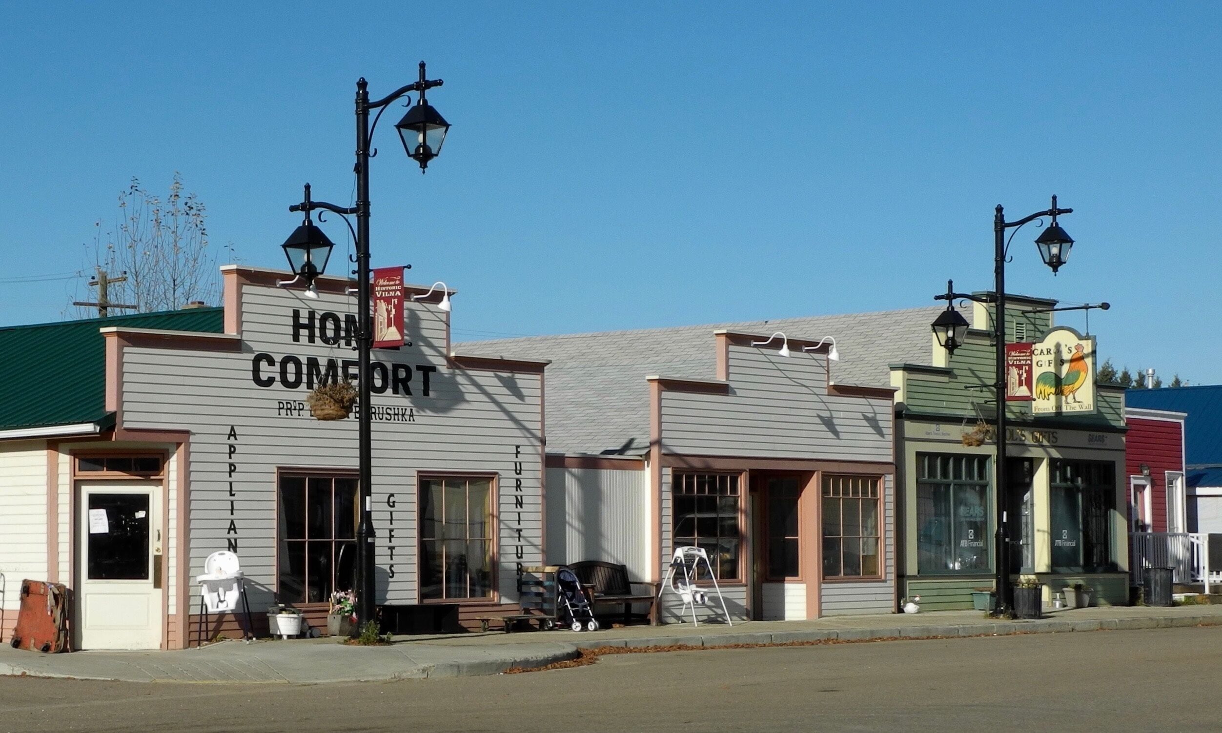A row of vintage storefronts with signs for 'Home Comfort' and 'Carson's Gifts'.