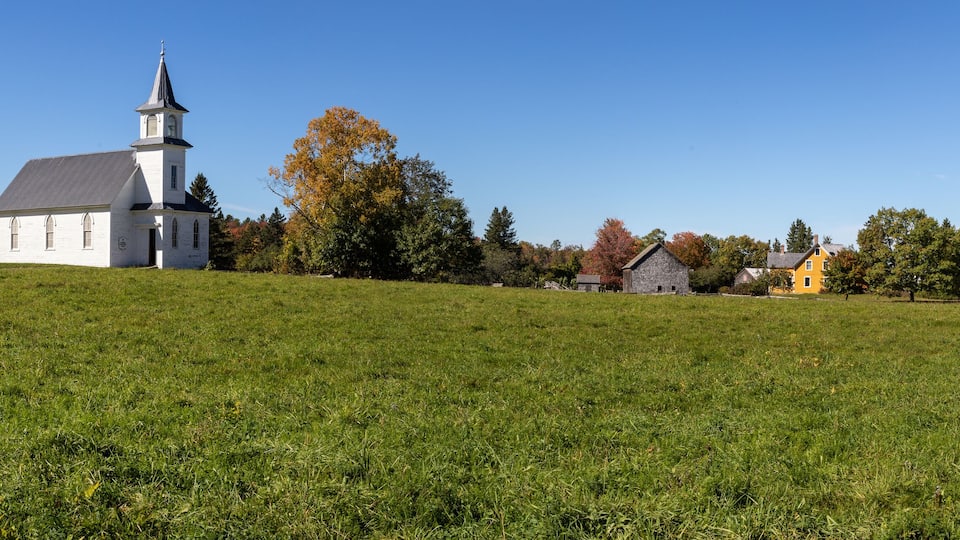 A white church with a steeple, a yellow house, and a barn on a grassy hill.