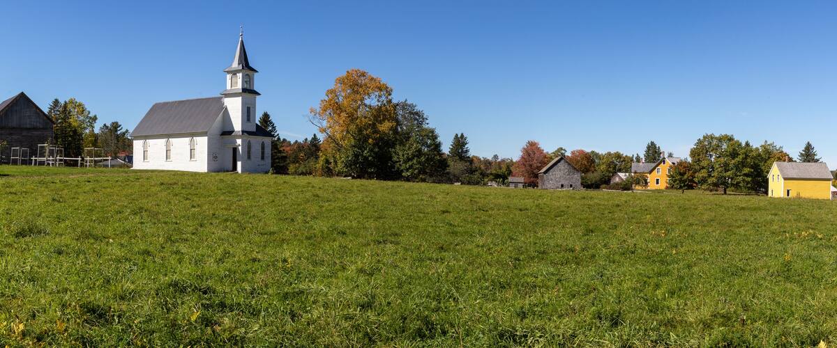 A white church with a steeple, a yellow house, and a barn on a grassy hill.