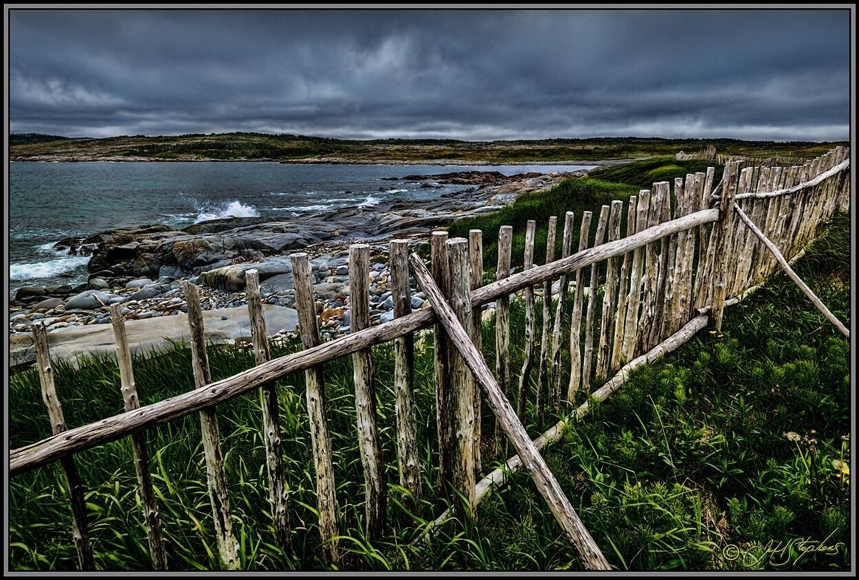 A wooden fence along a rocky shoreline with waves crashing onto the beach and a cloudy sky above.
