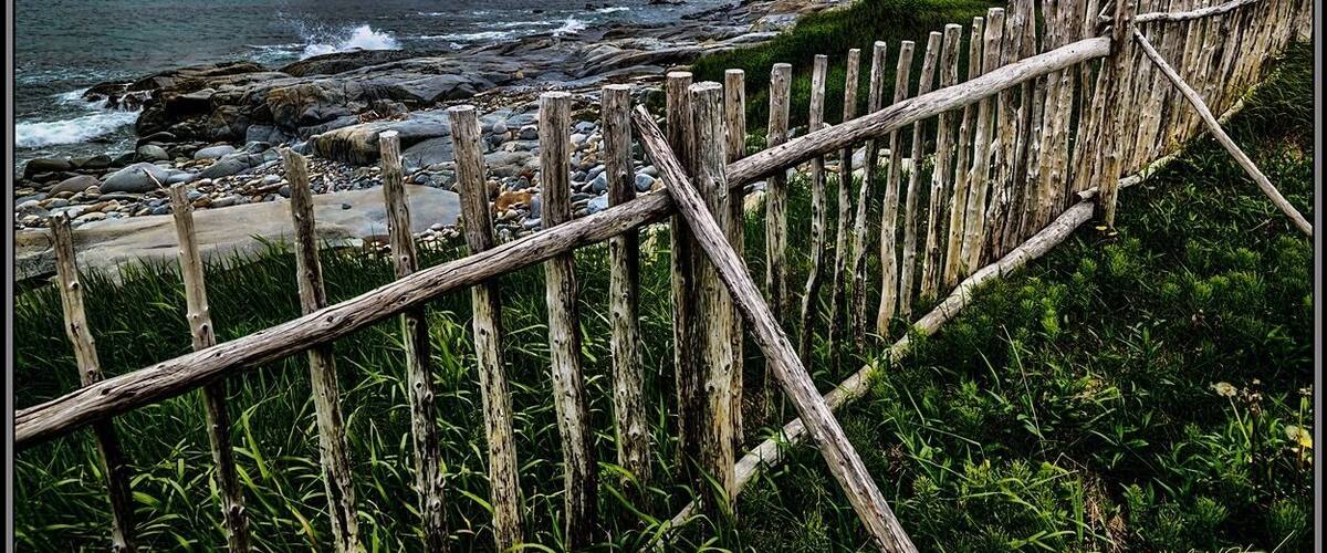 A wooden fence along a rocky shoreline with waves crashing onto the beach and a cloudy sky above.