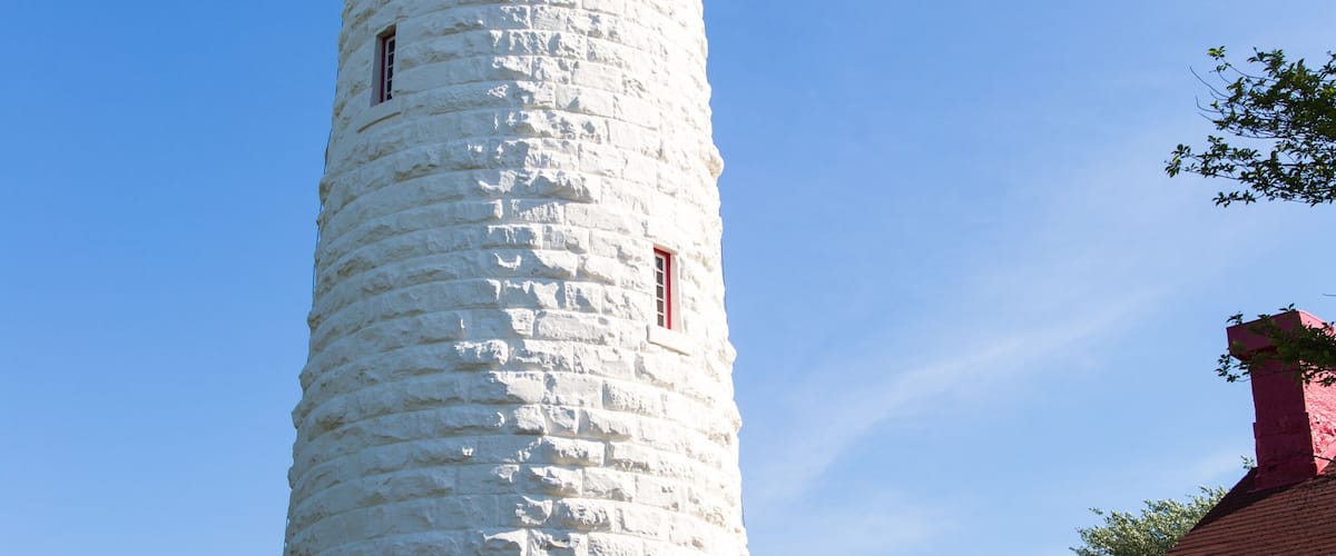 A tall white lighthouse with a red top, surrounded by greenery and a small building with a red roof.
