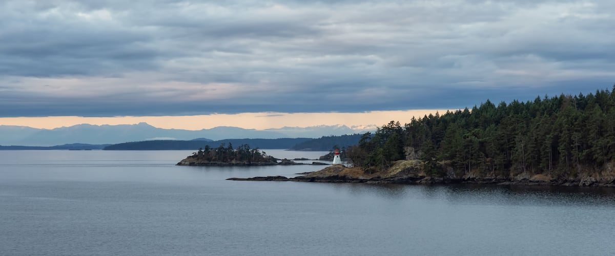 A calm lake with a lighthouse on a small island, surrounded by dense forest and distant mountains under a cloudy sky.