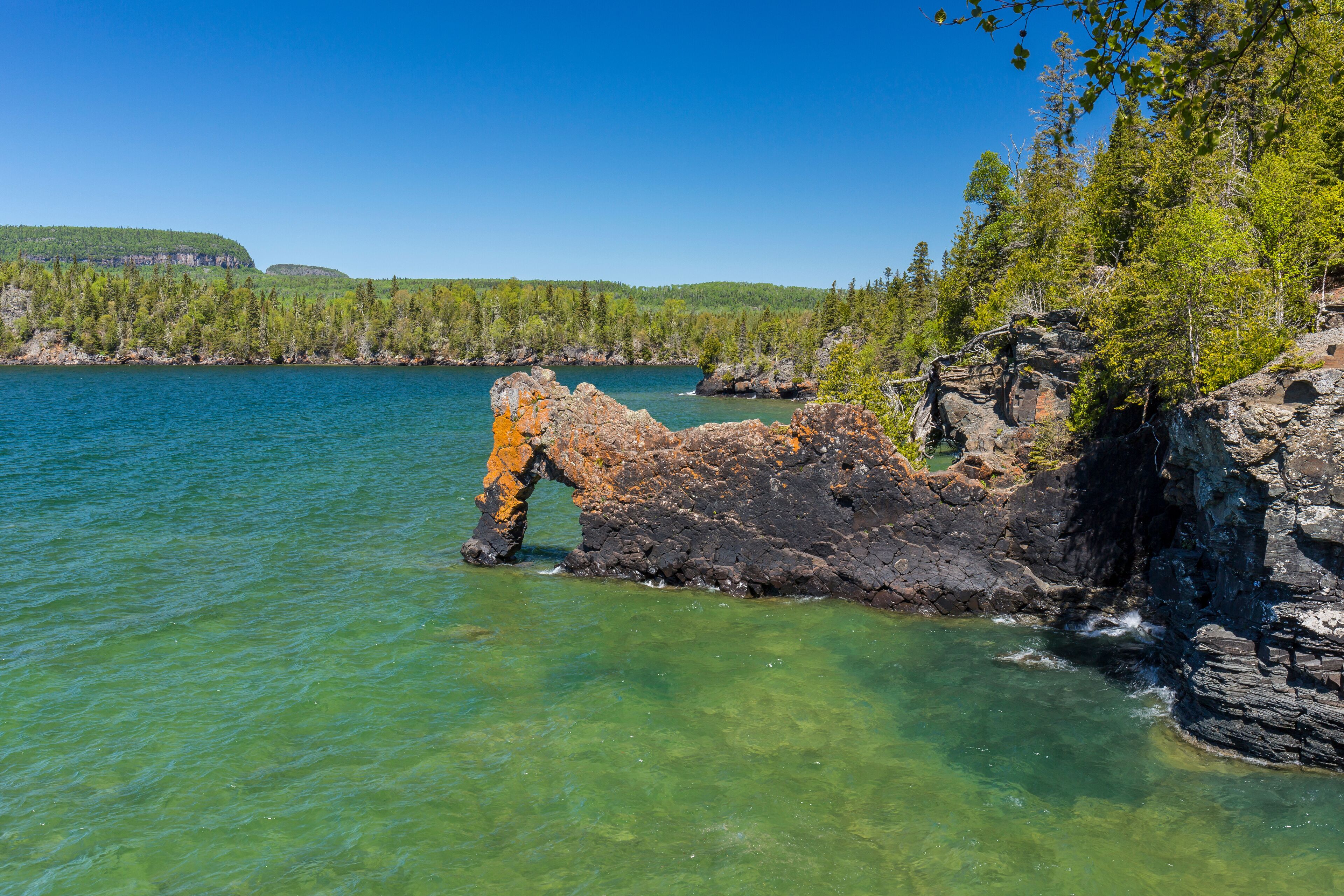 A rocky shoreline with clear water, surrounded by dense green forest and a distant hill.