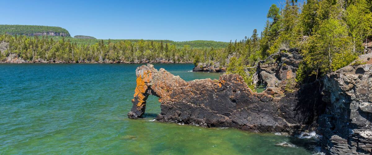 A rocky shoreline with clear water, surrounded by dense green forest and a distant hill.