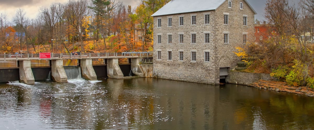 A stone building with a metal roof, a bridge over a river, and autumn-colored trees.